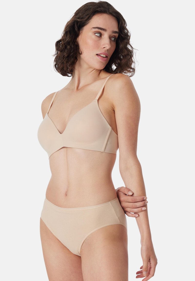 Femme portant un soutien-gorge nude sans coutures et un slip taille haute assorti, debout avec le bras plié et regardant sur le côté, sur fond blanc.