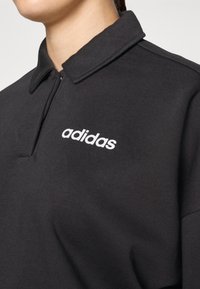Melns polo krekls ar rāvējslēdzēju, uz krūtīm ar baltu "adidas" logo. Izgatavots no gludas, teksturētas auduma ar klasisku apkakles dizainu.
