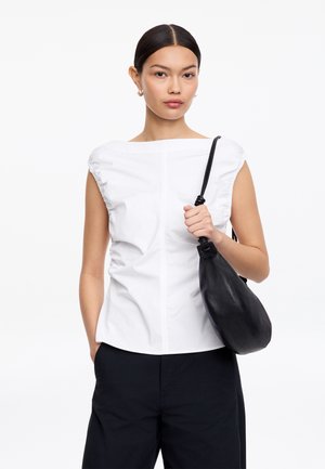 Frau mit dunklen Haaren, die ein weißes ärmelloses Oberteil und schwarze Hosen trägt, hält eine schwarze Lederschultertasche vor einem schlichten weißen Hintergrund.