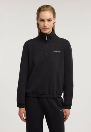 Kobieta ubrana w czarną bluzę zapinaną na zamek oraz pasujące czarne spodnie z logo "Trussardi 1911" na lewej piersi i lewym udzie.