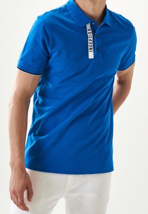 Polo bleu en coton, avec un col, trois boutons et une étiquette blanche contrastante portant "INCEPTION" sur le patte de boutonnage.