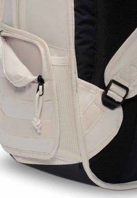 Κοντινό πλάνο πλαϊνής τσέπης μπεζ σακιδίου με φερμουάρ και ρυθμιζόμενο λουρί σε μαύρο υφασμάτινο φόντο.