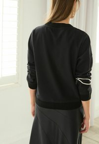 Pull-over noir avec col et poignets côtelés, orné de rayures blanches sur les manches. Porté sur une jupe noire plissée.