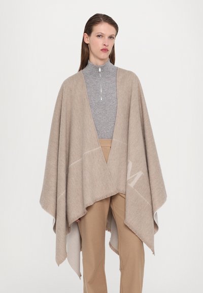 Beiger Poncho aus weichem Material mit drapiertem Design und fransigen Kanten. Kombiniert mit einem grauen Oberteil mit hohem Kragen und beigen Hosen.