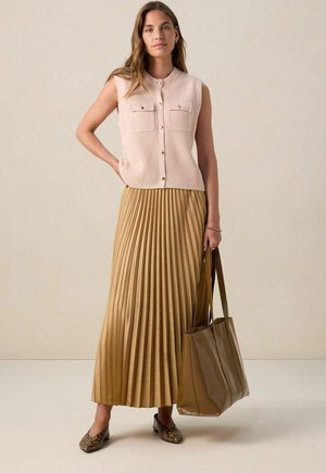Top sans manches côtelé rose clair avec deux poches, associé à une jupe maxi plissée dorée. Sac fourre-tout marron et chaussures plates à motifs.