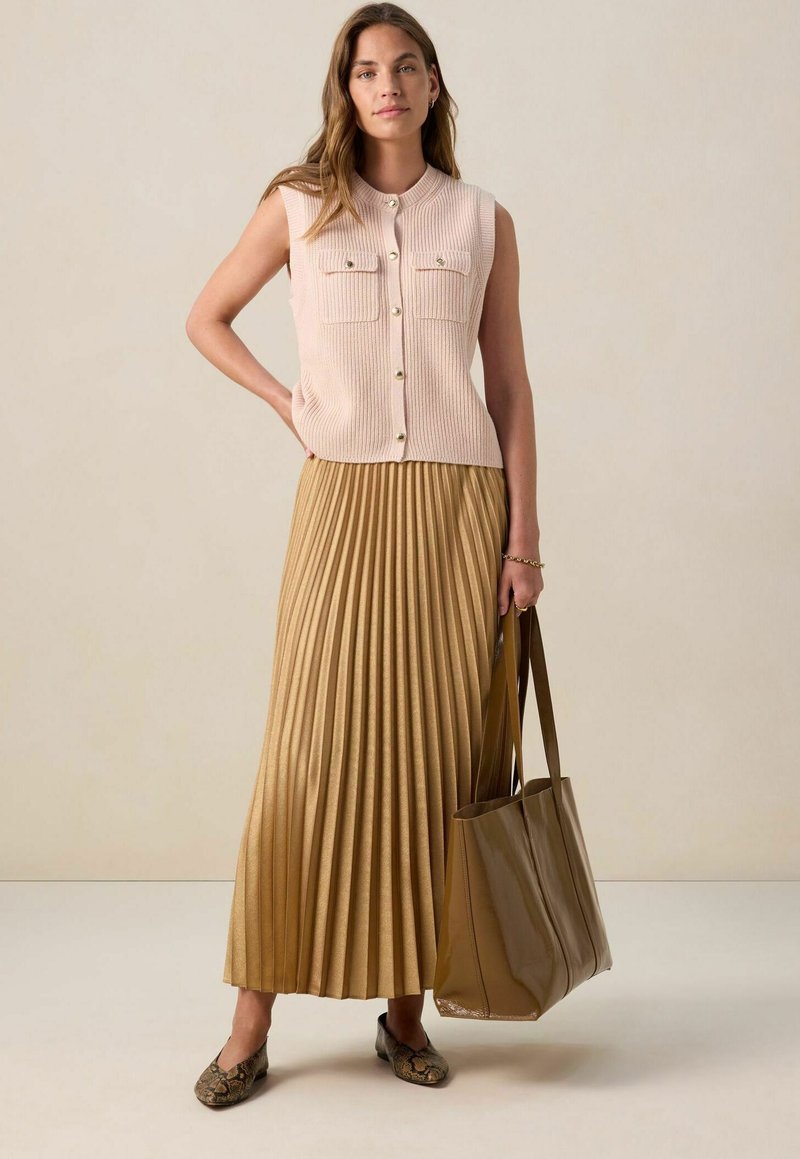 Top sans manches côtelé rose clair avec deux poches, associé à une jupe maxi plissée dorée. Sac fourre-tout marron et chaussures plates à motifs.