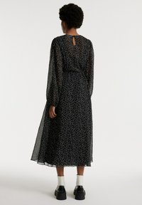 Zwarte jurk met stippen, lange doorzichtige mouwen, een sleutelgatdetail op de rug en een vloeiende silhouet, gecombineerd met stevige zwarte schoenen en witte sokken.