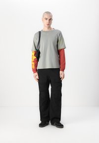 T-shirt grigio a maniche corte sovrapposto a una maglietta rossa a maniche lunghe, abbinato a pantaloni cargo neri e scarpe da ginnastica nere. Elementi di design minimal.