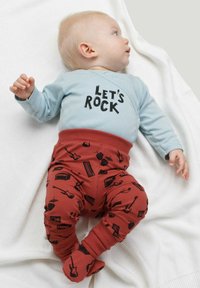 Camicia a maniche lunghe azzurro chiaro con la scritta "Let's Rock", abbinata a pantaloni rossi con un motivo di strumenti musicali neri, su una coperta bianca testurizzata.