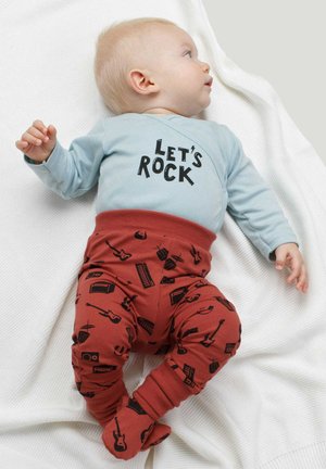 Jasnoniebieska koszula z długim rękawem z napisem "Let's Rock", w połączeniu z czerwonymi spodniami w czarny wzór muzycznych instrumentów, na fakturowanym białym kocu.