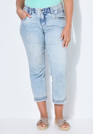 Kobieta w jasnoniebieskich krótkich dżinsach z haftowanym brzegiem i beżowych sandałach wsuwanych, stojąca na białym tle.