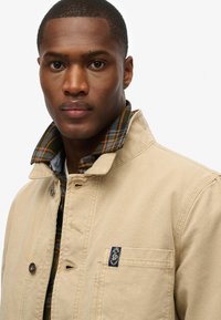 Beige Canvas-Jacke mit Druckknopfverschluss und kariertem Futter in Orange und Grün. Mit einer Brusttasche und einem markierten Patch.