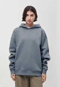 Jeune femme aux cheveux courts et foncés portant un sweat à capuche gris LeGer et un pantalon beige, se tenant devant un fond clair, face à l'objectif.