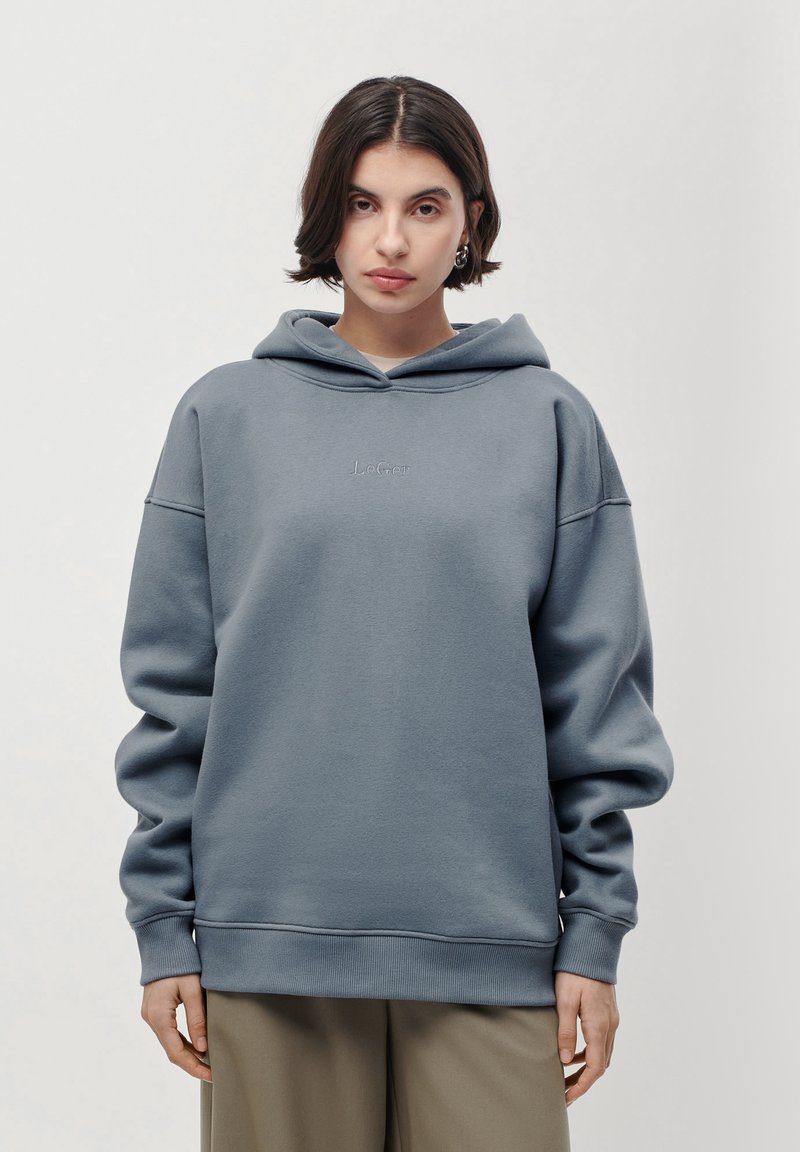 Jeune femme aux cheveux courts et foncés portant un sweat à capuche gris LeGer et un pantalon beige, se tenant devant un fond clair, face à l'objectif.