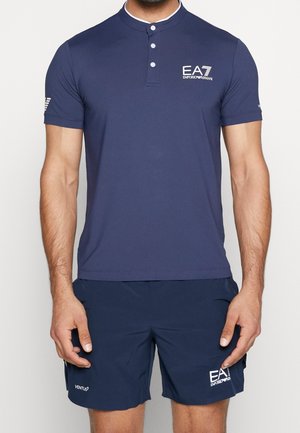Mann trägt navyblaues EA7 Emporio Armani kurzärmeliges Polohemd und passende Shorts mit Logos auf Brust und Bein.