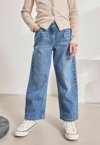 Široké modré džíny z denimu s opotřebovaným vzhledem, vysokým pasem a nedokončeným lemem. V kombinaci s bílými teniskami.