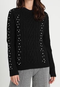 Czarny sweter wykonany na drutach z teksturowanym wzorem w warkocze i białymi perłowymi detalami wzdłuż rękawów. Żebrowany dekolt i mankiety.