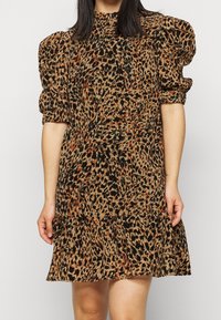 Eläinkuosilla varustettu mekko, jossa on puffihihat, korkea kaula-aukko ja levenevä hame. Värit ovat beige, musta ja ruskea oransseilla yksityiskohdilla.