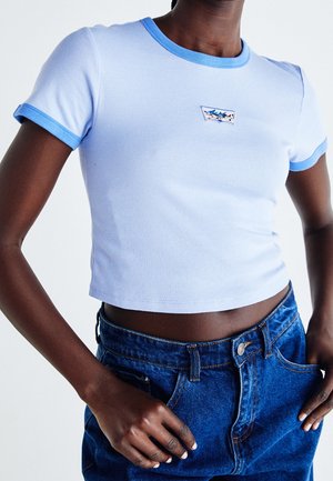 Persoon draagt een lichtblauw cropped t-shirt met donkerblauwe bies en blauwe spijkerbroek, handen rusten nabij de zakken.
