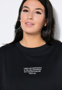 Sort t-shirt med rund halsudskæring og korte ærmer, der har hvid tekst: "JEG KLÆDER MIG FOR KOMPLIMENTER I PIGEBADRUMMET, IKKE FOR DRENGE, DET ER SIKKERT. TRO MIG."
