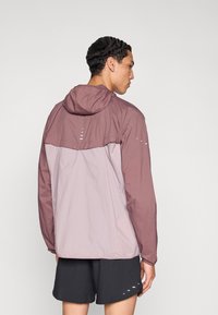 Kerge kahetooniline tuulejope lavendlivärvi ja kahvatu roosa toonis, kapuutsiga, millel on helendavad detailid ja elastsed mansetid, näidatud tagantpoolt.