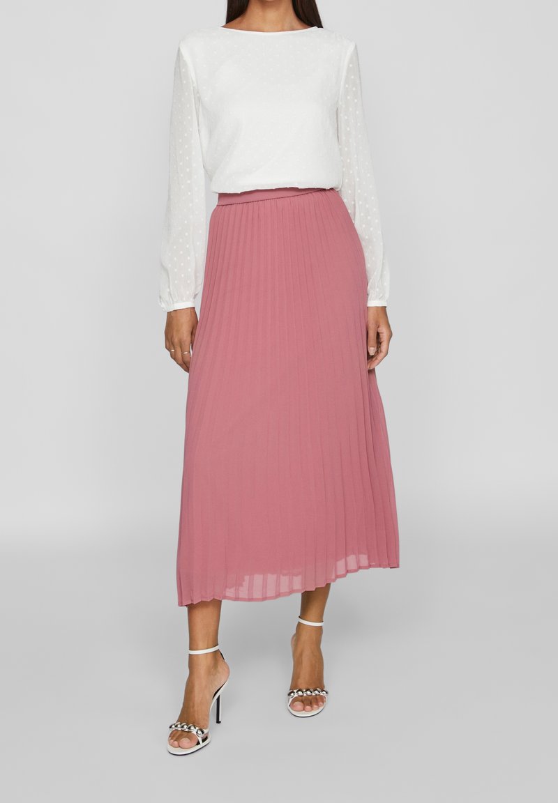 Femme portant un chemisier blanc à manches longues transparentes à pois et une jupe midi plissée rose taille haute avec des talons blancs.