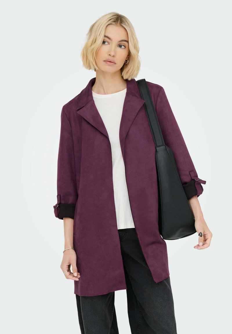 Fialový zamietaný trench coat s výstrihom, zrolovanými rukávmi a voľným strihom; doplnený čiernou kabelkou a bielym topom.