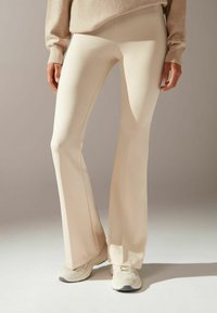 Beige utsvängda yogabyxor tillverkade av elastiskt tyg, med en slät textur och en slim fit genom låren, kombinerat med ljusa sneakers.