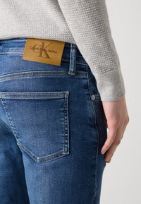 Tamne plave traperice glatke teksture, s bež kožnom etiketom na struku koja nosi logotip "Calvin Klein Jeans" i standardnim džepovima.