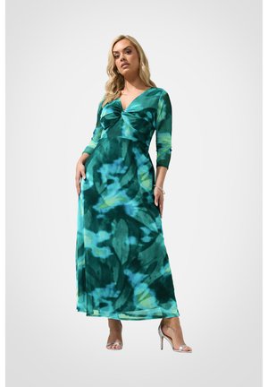 Kobieta w długiej, zielono-niebieskiej sukience tie-dye z rękawami do łokcia i srebrnych sandałach na wysokim obcasie, stojąca na białym tle.