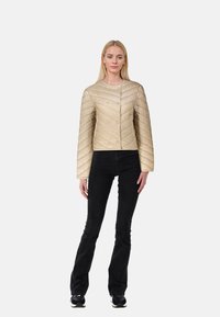 Beige vadderad kort jacka med dubbelknäppt framtill, långa ärmar och zigzag-mönster, bärs över svarta utsvängda byxor och sneakers.