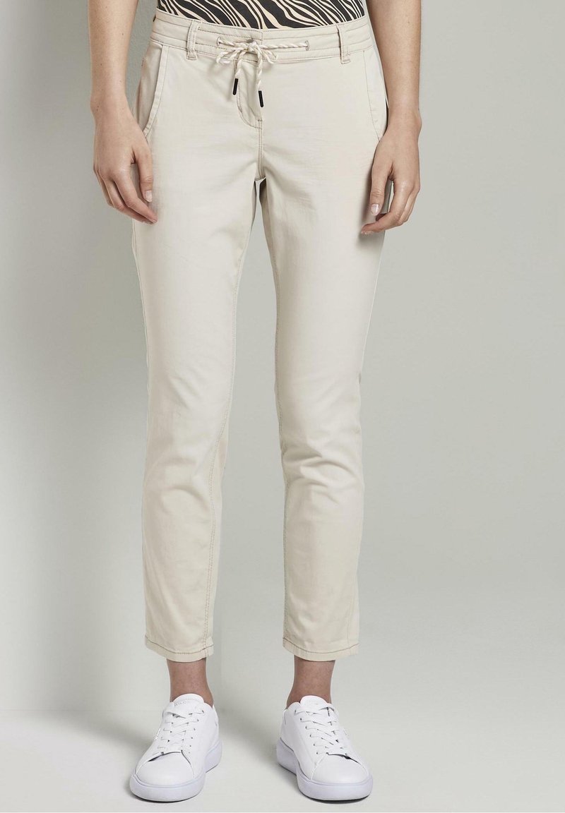Beige Baumwollmischungshosen mit einem Kordelzugbund, schmal zulaufenden Beinen und Seitentaschen, kombiniert mit weißen Sneakers. Einfaches Design, minimale Details.