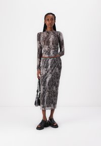 Svart og grå slangeskinnmønster topp med lange ermer og høy hals, sammen med et matchende skjørt. Modellen holder en svart veske og har på seg platåsandaler.