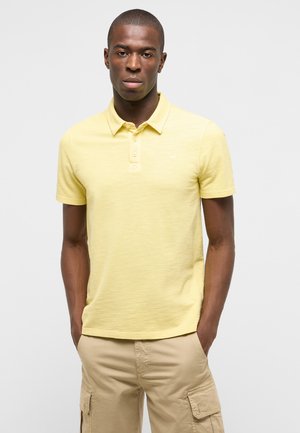 Mann trägt ein hellgelbes kurzärmeliges Poloshirt und beige Cargo-Shorts, steht mit den Händen in den Taschen vor einem einfarbigen Hintergrund.