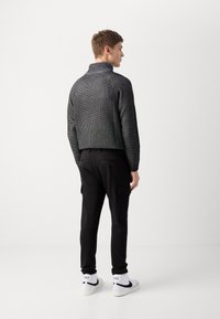 Svart strikket genser med høy krage, kombinert med svarte joggebukser med sidelommer. Hvite sneakers med svarte detaljer.
