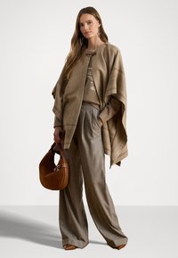 Poncho beige con chiusura a bottone, indossato sopra un leggero maglione, abbinato a pantaloni grigi a gamba larga e una borsa marrone.