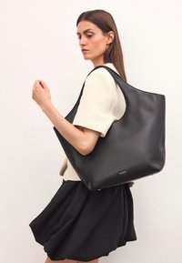 Svart läder tote-väska med en slät yta, breda axelremmar och logomarkeringar. Modellen bär en krämfärgad topp och svart kjol.