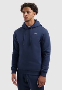 Jongeman staat in een marineblauwe hoodie met "BALLIN" logo op de borst en bijpassende marineblauwe joggingbroek tegen een effen achtergrond.