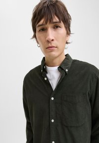 Mørkegrønn kordfl�øyelskjorte med knappet krage, enkelt brystlomme og hvite knapper, lagd over en hvit T-skjorte.