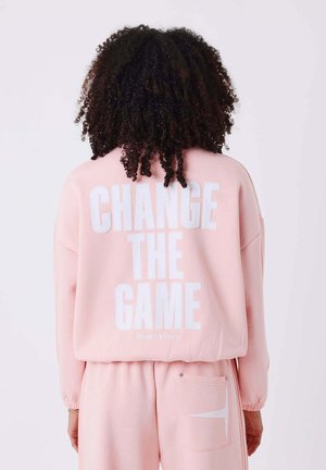 Personne aux cheveux bouclés portant un sweat-shirt et un pantalon roses, avec le texte "CHANGE THE GAME" au dos du sweat-shirt.