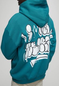 Persona che indossa un hoodie turchese con un audace design in graffito bianco e nero sulla schiena, girata di spalle su uno sfondo semplice.