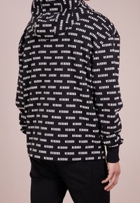 Person som bär en svart huvtröja med ett vitt upprepat "VERSUS" textmönster, i kombination med svarta byxor, stående mot en enfärgad bakgrund.