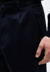 Κοντινό πλάνο του δεξιού χεριού ενός ατόμου που ακουμπά στο πλάι του, φοράει σκούρο μπλε παντελόνι με πλαϊνή τσέπη και λεπτομέρεια πιέτας.