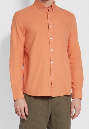 Homme portant une chemise orange à manches longues, boutonnée, avec un col classique et un pantalon sombre texturé sur un fond uni.