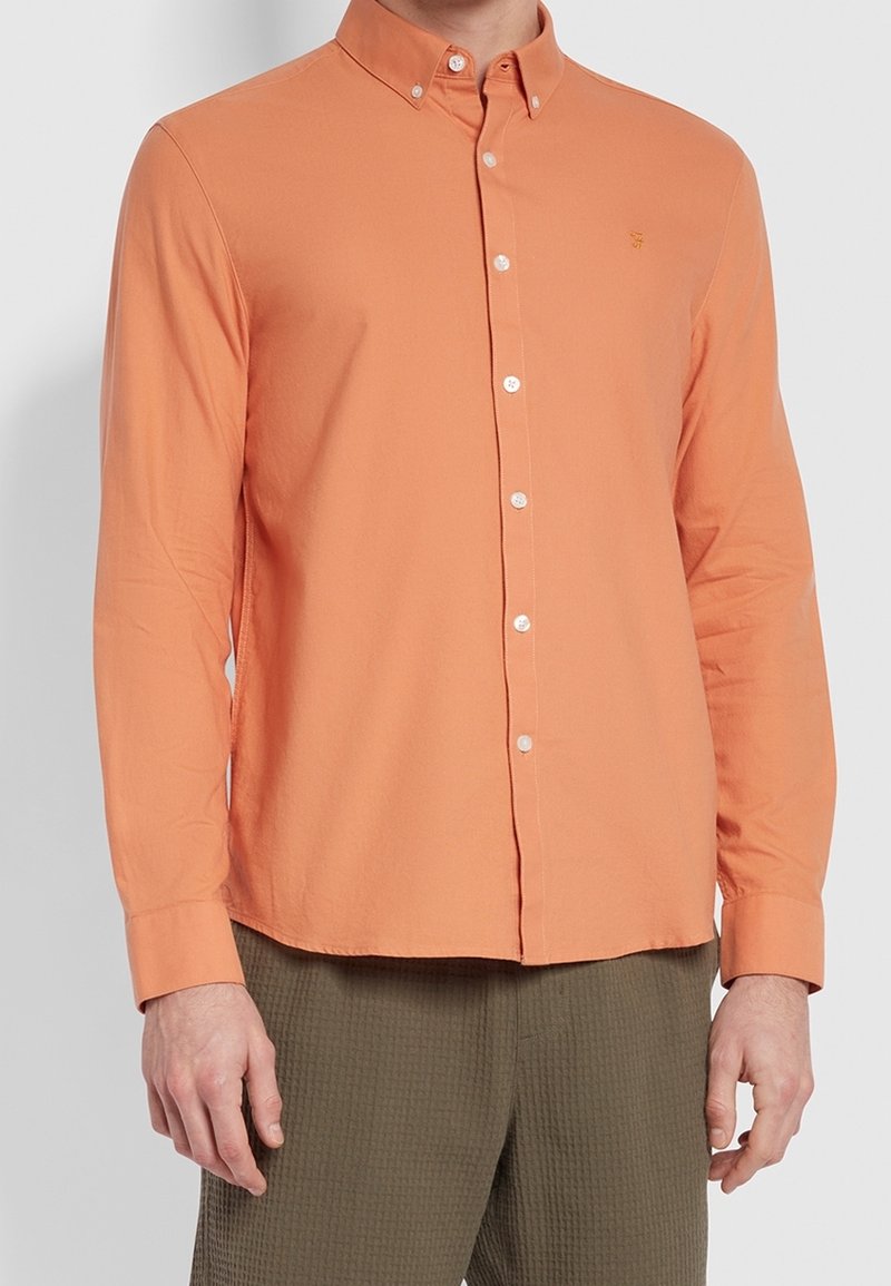 Homme portant une chemise orange à manches longues, boutonnée, avec un col classique et un pantalon sombre texturé sur un fond uni.