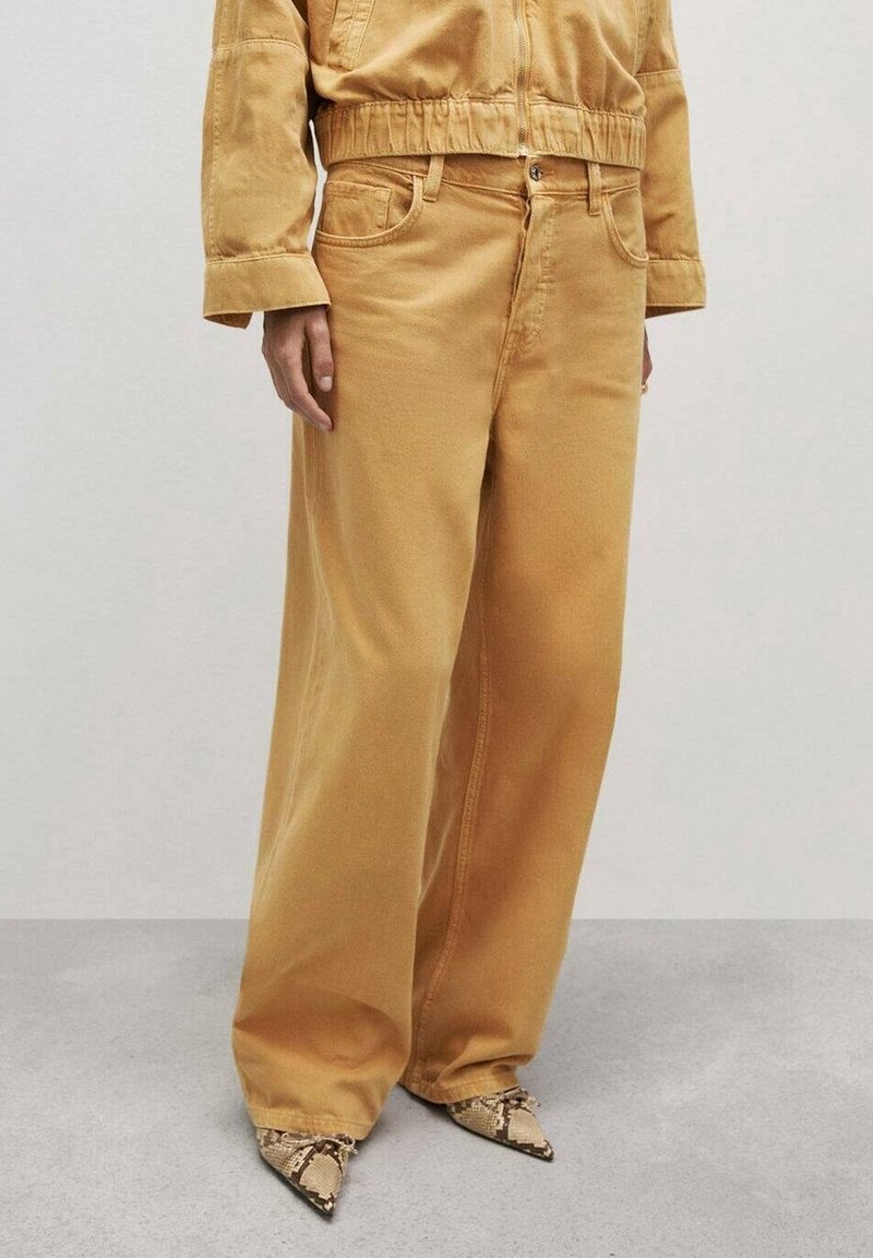 Pantalon large jaune moutarde associé à des chaussures pointues en cuir effet peau de serpent, et une veste courte assortie à manches longues.