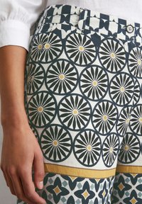 Pantalons à motifs présentant des motifs circulaires en noir, blanc et jaune, avec un design géométrique en bleu et blanc à l'ourlet, fabriqués en tissu léger.