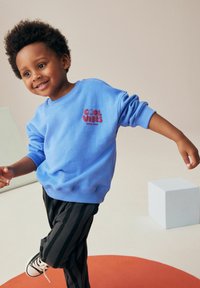 Sweatshirt bleu avec "COOL VIBES" en texte rouge, poignets et ourlet côtelés, associé à un pantalon rayé noir et des baskets blanches.