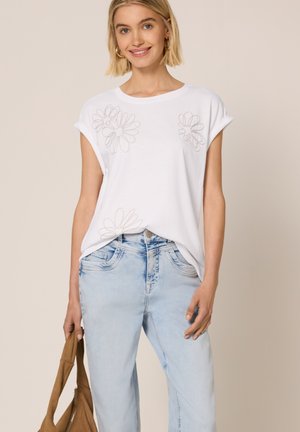 Młoda kobieta w białej koszulce z kwiatowym haftem i jasnoniebieskich dżinsach trzymająca brązową torbę, uśmiechająca się na beżowym tle.