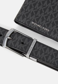 Set di cintura e portafoglio Michael Kors neri con motivo testurizzato del logo MK e dettagli in argento sul cinturino e sul logo, su sfondo bianco.